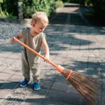 charming child sweeping concrete pavement with broomstick