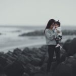 a woman in gray sweater and black pants standing while carrying her baby